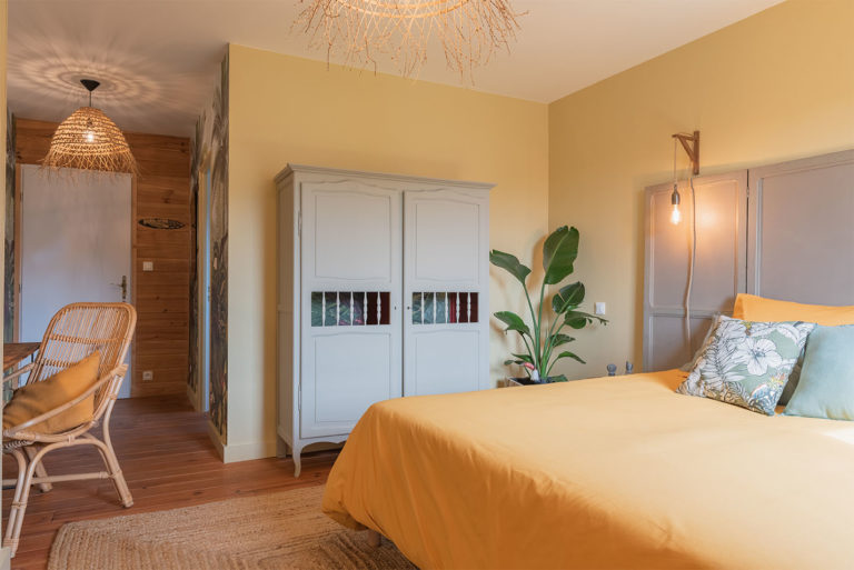 Photo d'un grand lit jaune avec armoire et vue sur l'entrÃ©e avec lustre en paille et plante verte.