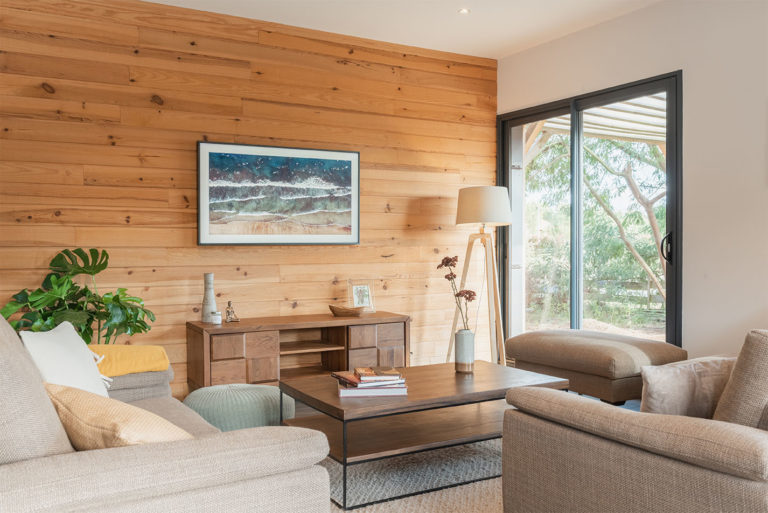 Photo d'un coin salon avec canapés, table basse et mobilier en bois devant une façade en bois avec une télévision masquée par une grande photographie de mer.