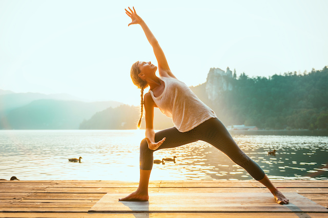 Photo d'une femme en posture de yoga main en l'air sur une terrasse en bois devant la montagne et un lac.