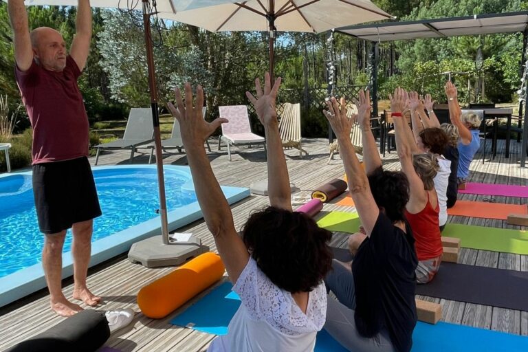 Photo d'un prof de yoga avec ses Ã©lÃ¨ves en postures mains levÃ©es devant une piscine.