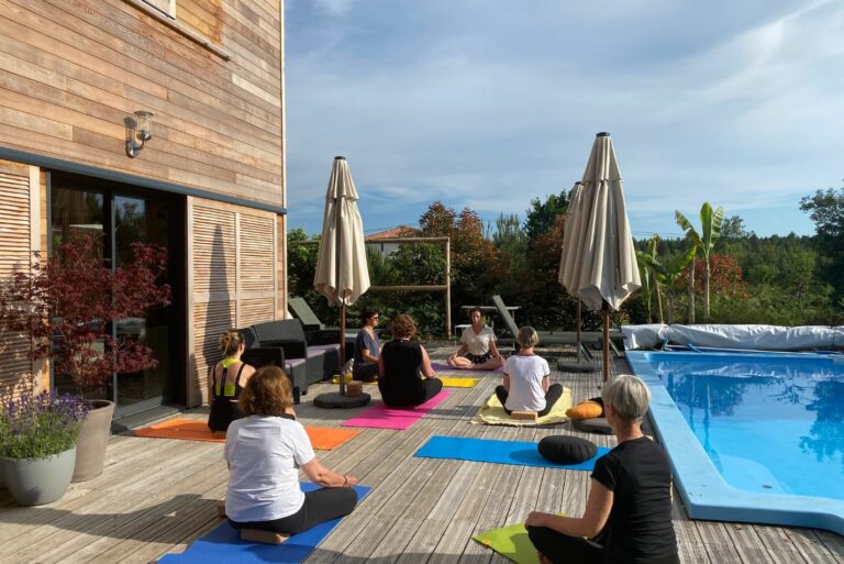 Photo d'un groupe de personnes en train de faire du yoga au bord de la piscine sur la terrasse en bois au soleil.