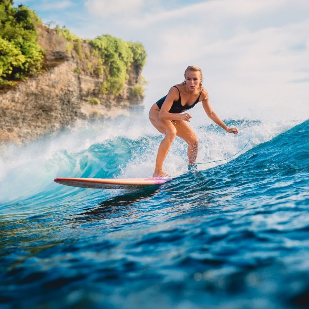 Photo d'une femme sur une planche de surf sur une vague bleue et fond de rocher et végétation.