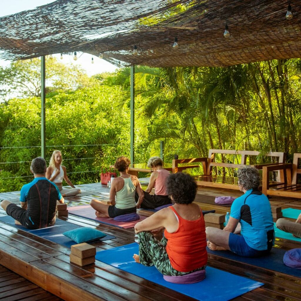 Photo d'un groupe de personnes en train de suivre un cours de yoga sur un deck en bois en extérieur.