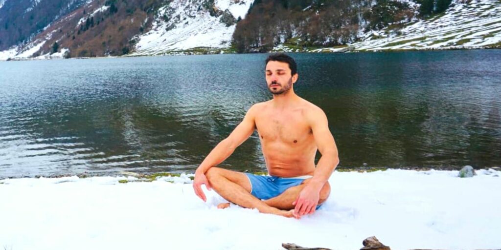 Photo d'un homme méditant torse nu sur la glace devant une étendue d'eau et des montagnes.