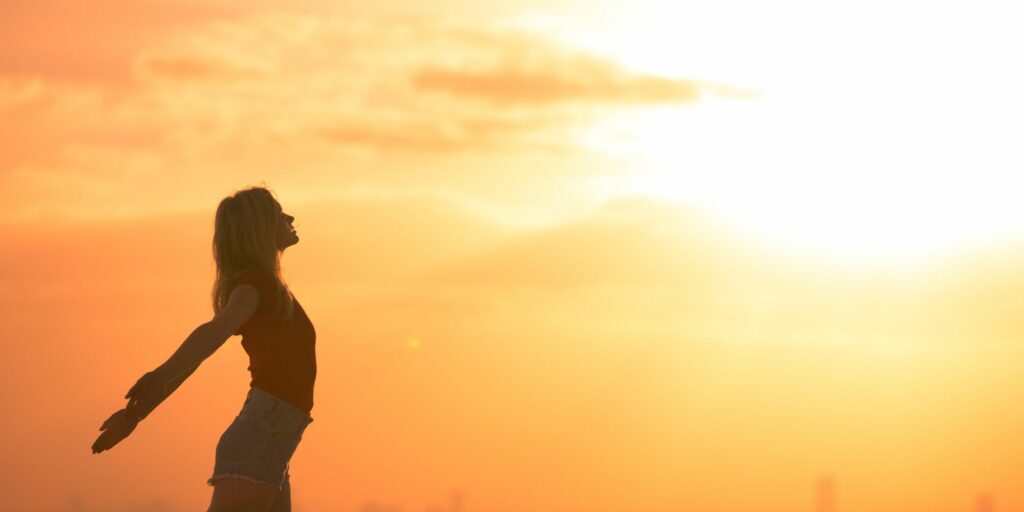 Photo d'une silhouette de femme tendant les bras en arrière sur un fond de ciel orangé par le soleil.